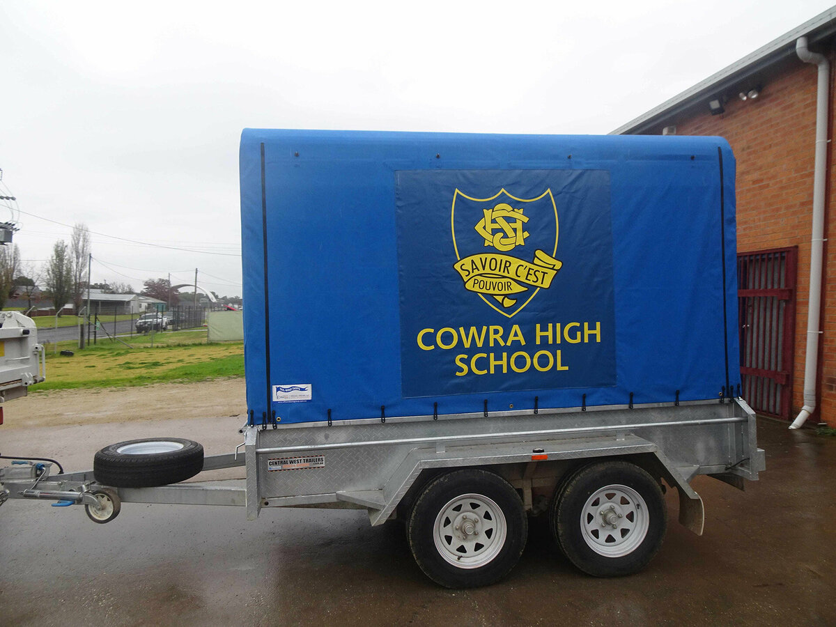 A trailer sitting in a driveway outside a brick building. The trailer has a blue canopy with the Cowra High School logo