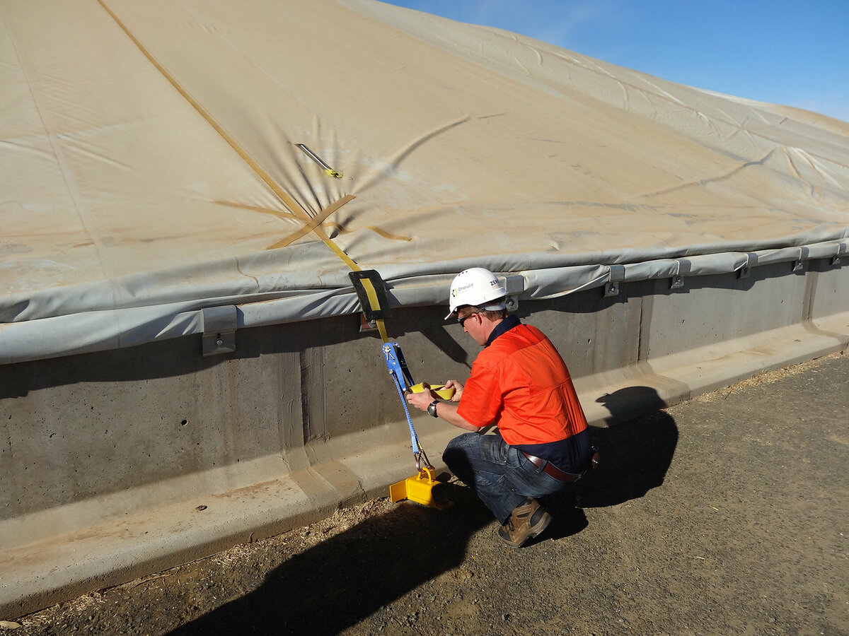 A man is wearing an orange shirt and white hard hat while fastening a GrainSafe cover strap