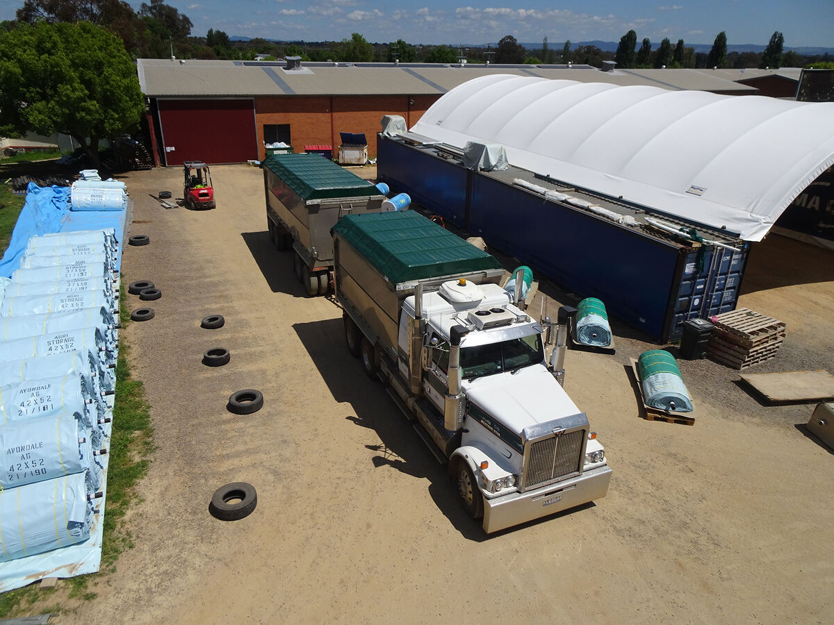 Aerial view of a truck parked in a dirt yard next to a shed. The truck has a retractable tarp on each trailer.