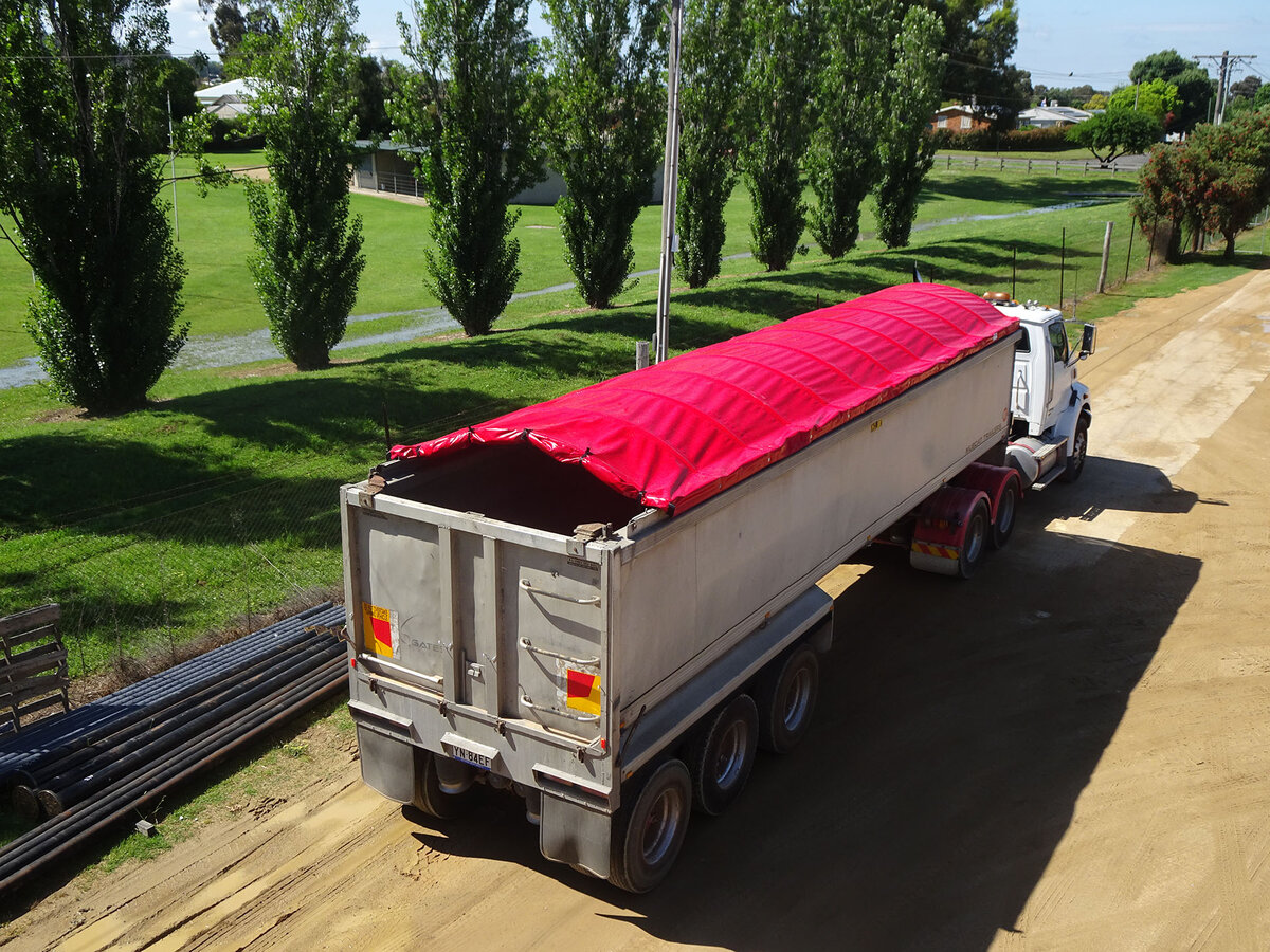 A truck drives down a dirt road lined with tall thin trees. The trailer has a red retractable tarp.
