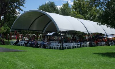 Large curved relocatable shade structure covers a large group of people sitting at tables in a park