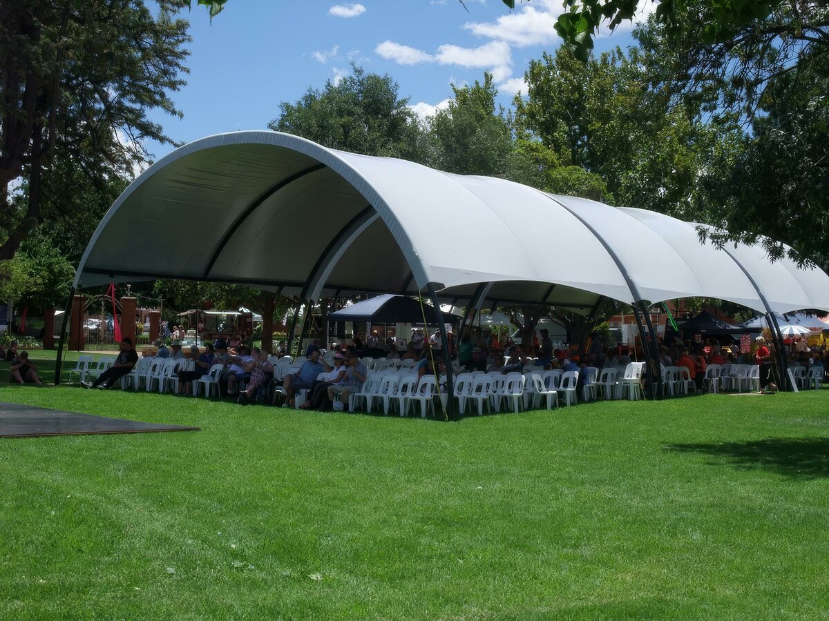 Large curved relocatable shade structure covers a large group of people sitting at tables in a park