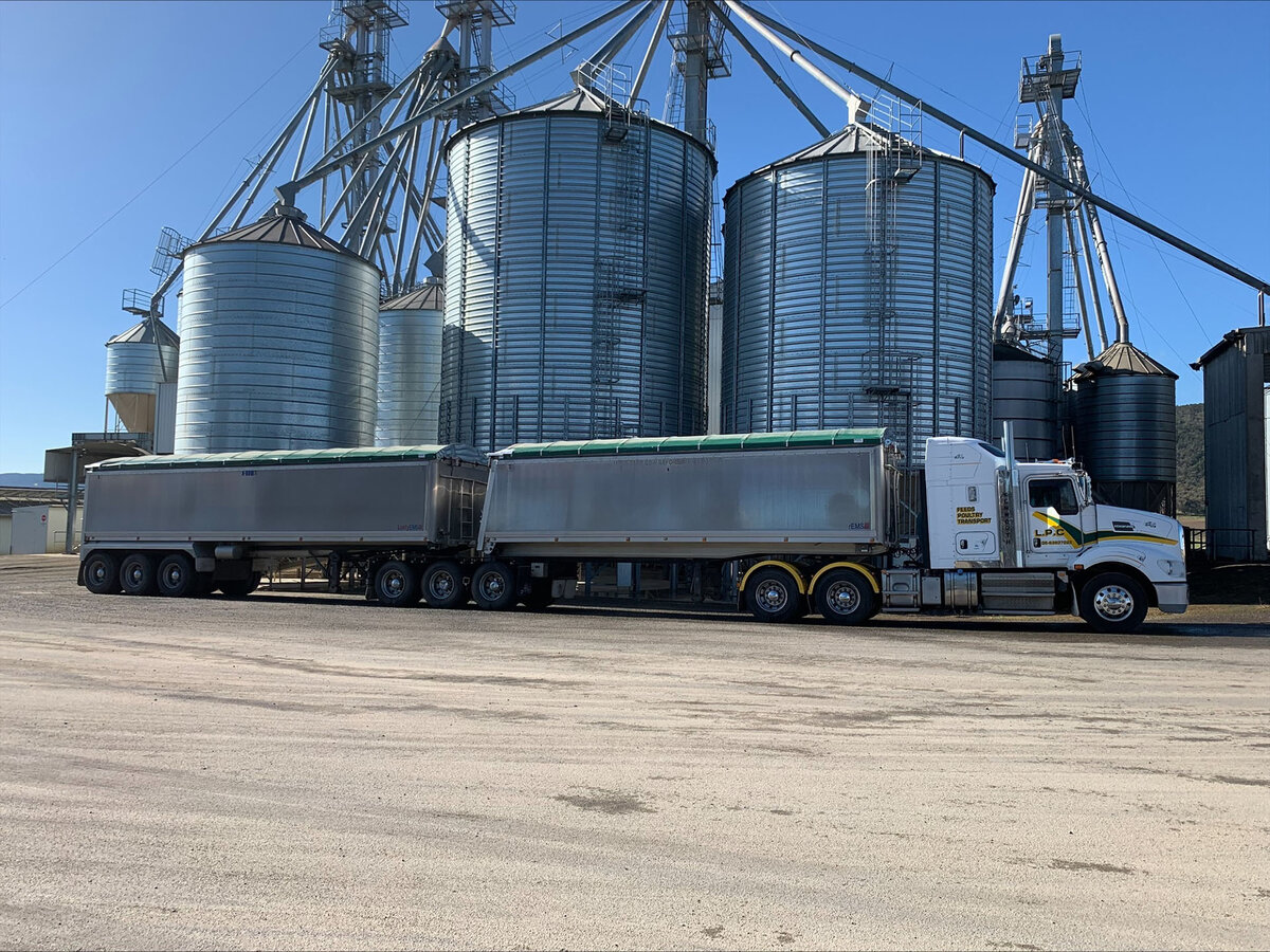 A truck and trailer is parked in front of several large grain silos