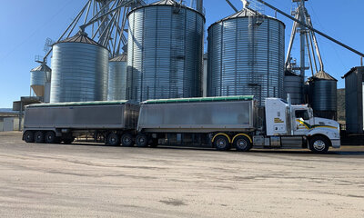 A truck and trailer is parked in front of several large grain silos